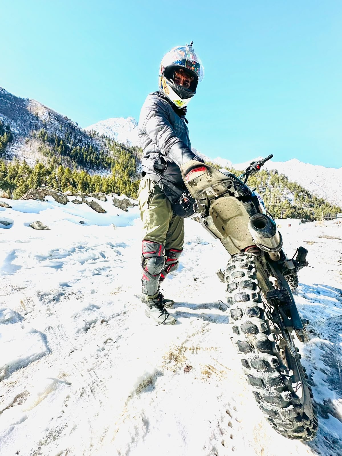 A rider in helmet and knee guards with his adventure bike on a snowfield below the mountains