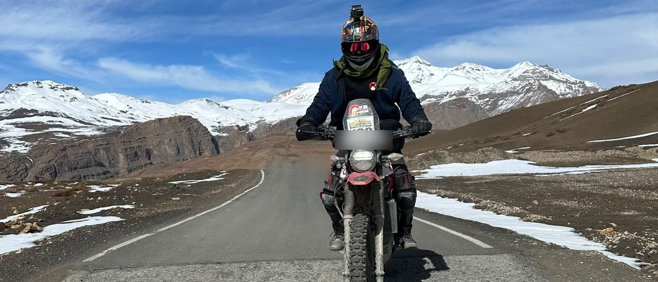 A rider on a dirt-streaked adventure bike on a high Spiti road below snow peaks